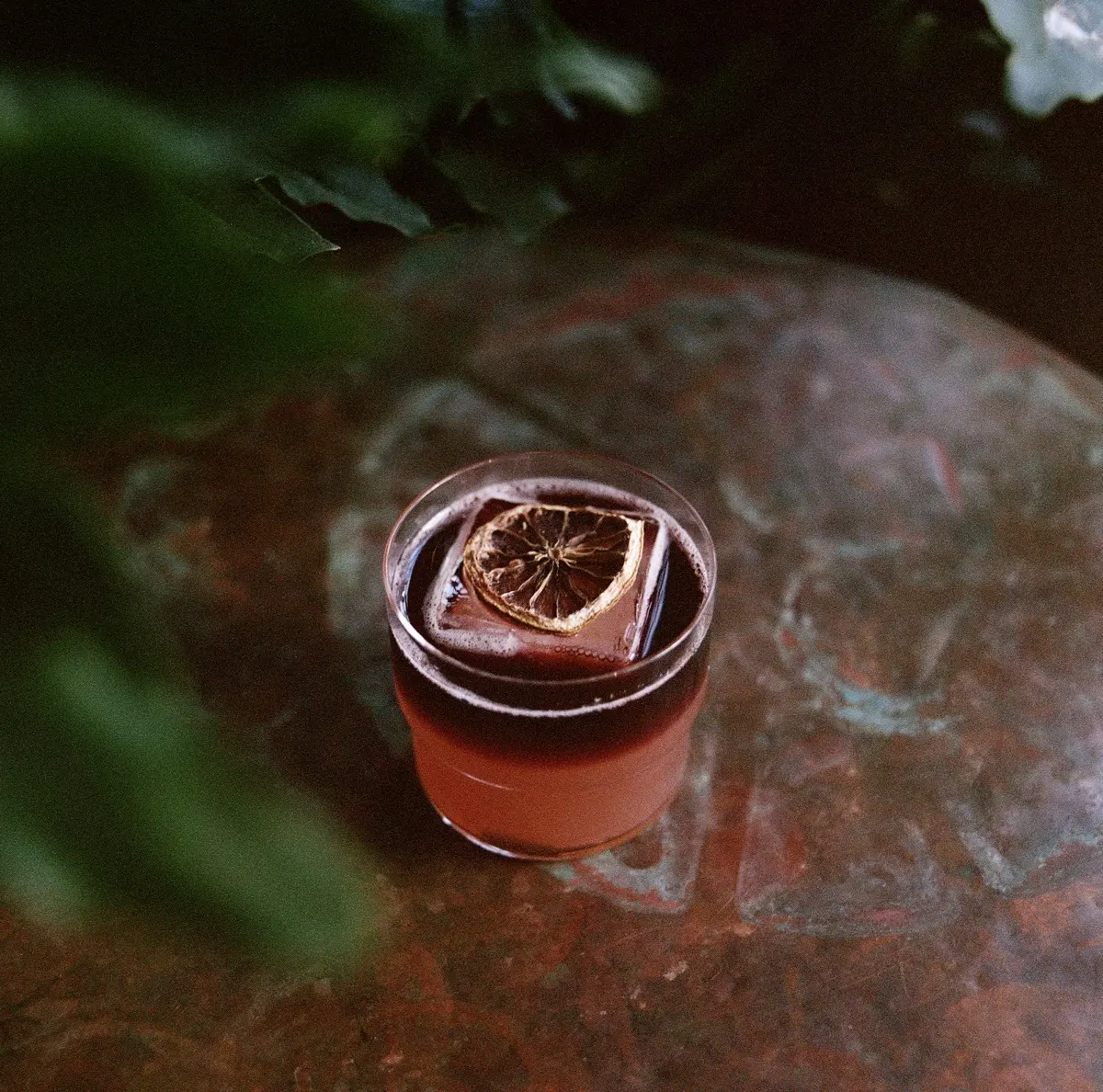 A dark cocktail on copper with a leaf shadow across it